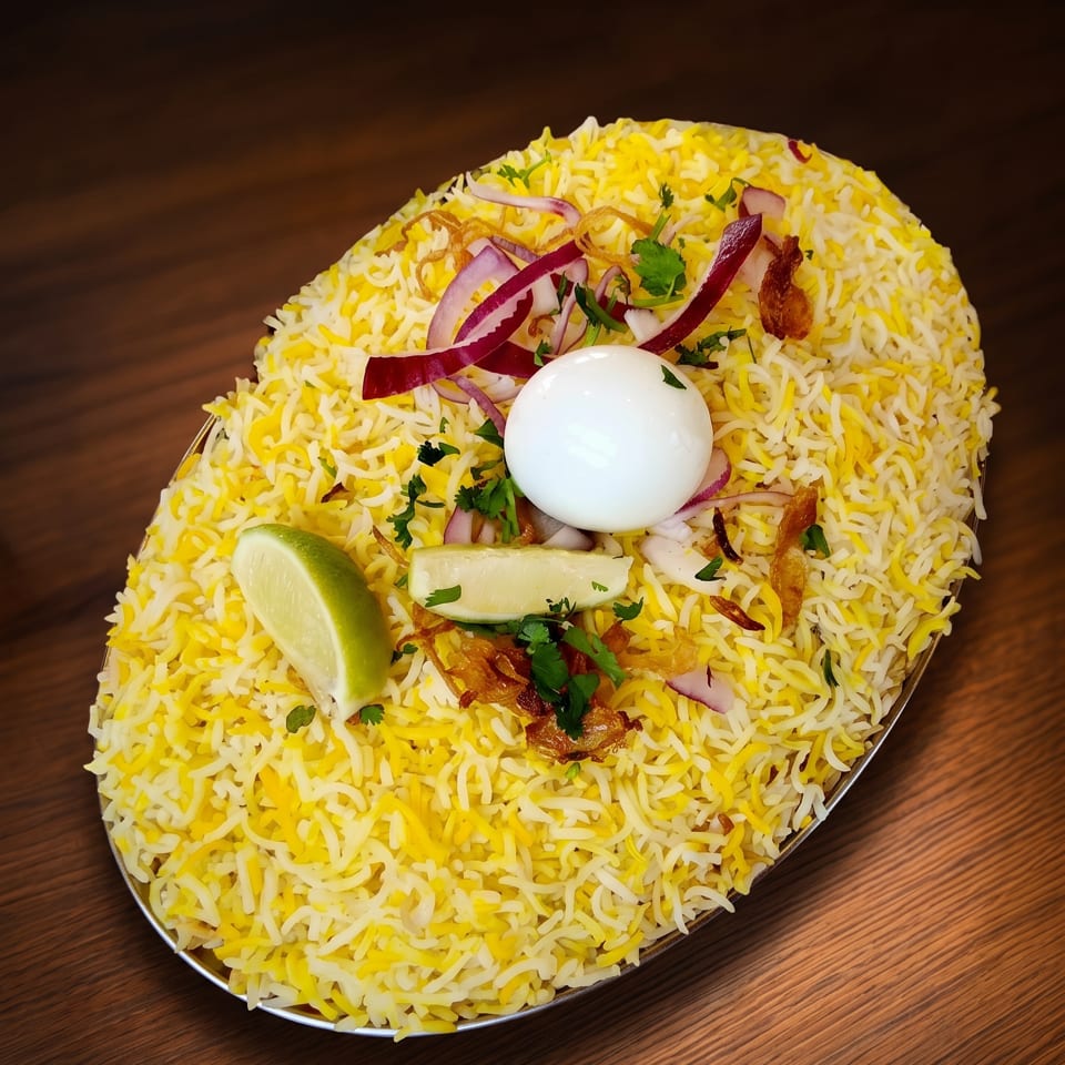 Oval plate of biryani with a boiled egg, red onions, cilantro, and lime on a wooden table.