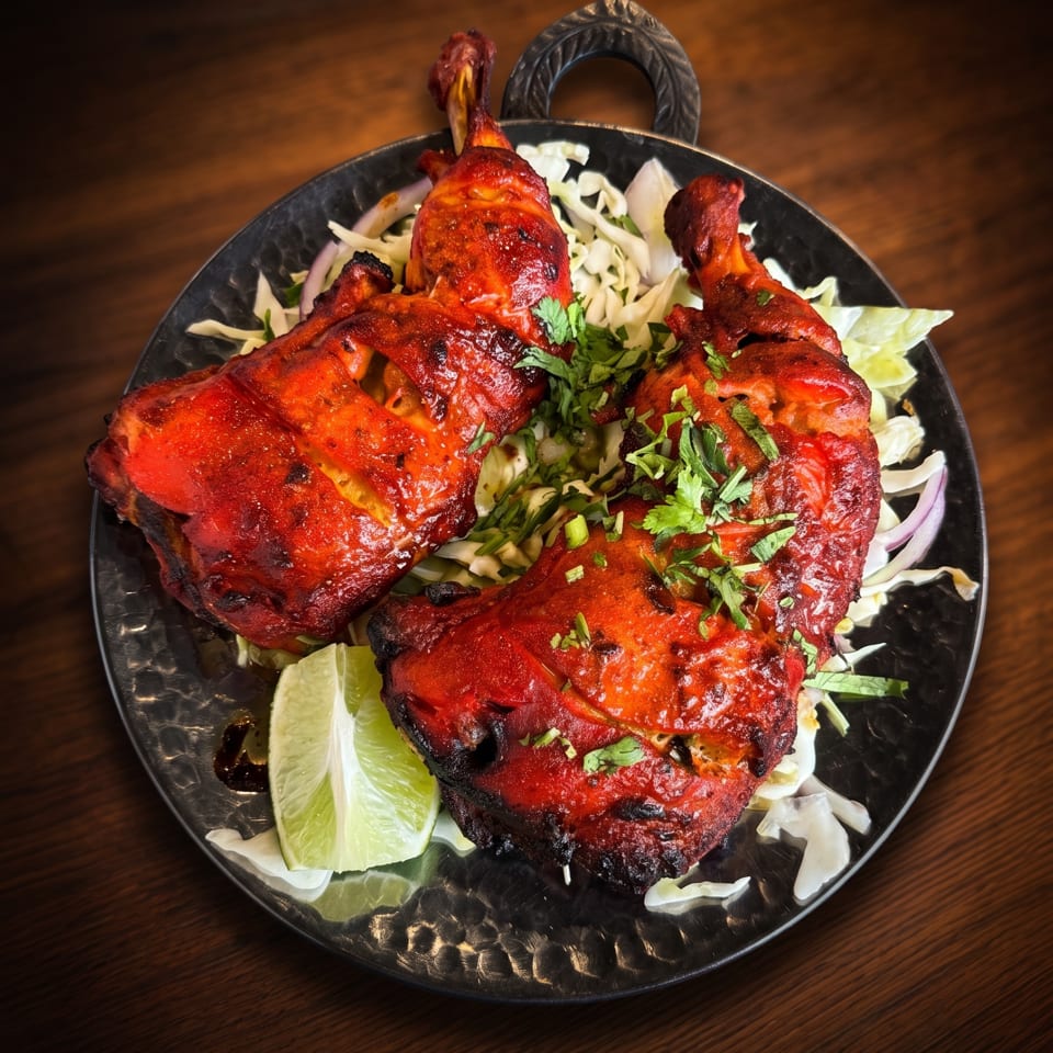Red tandoori chicken pieces on a bed of cabbage and onions, garnished with cilantro and lime, on a dark plate.