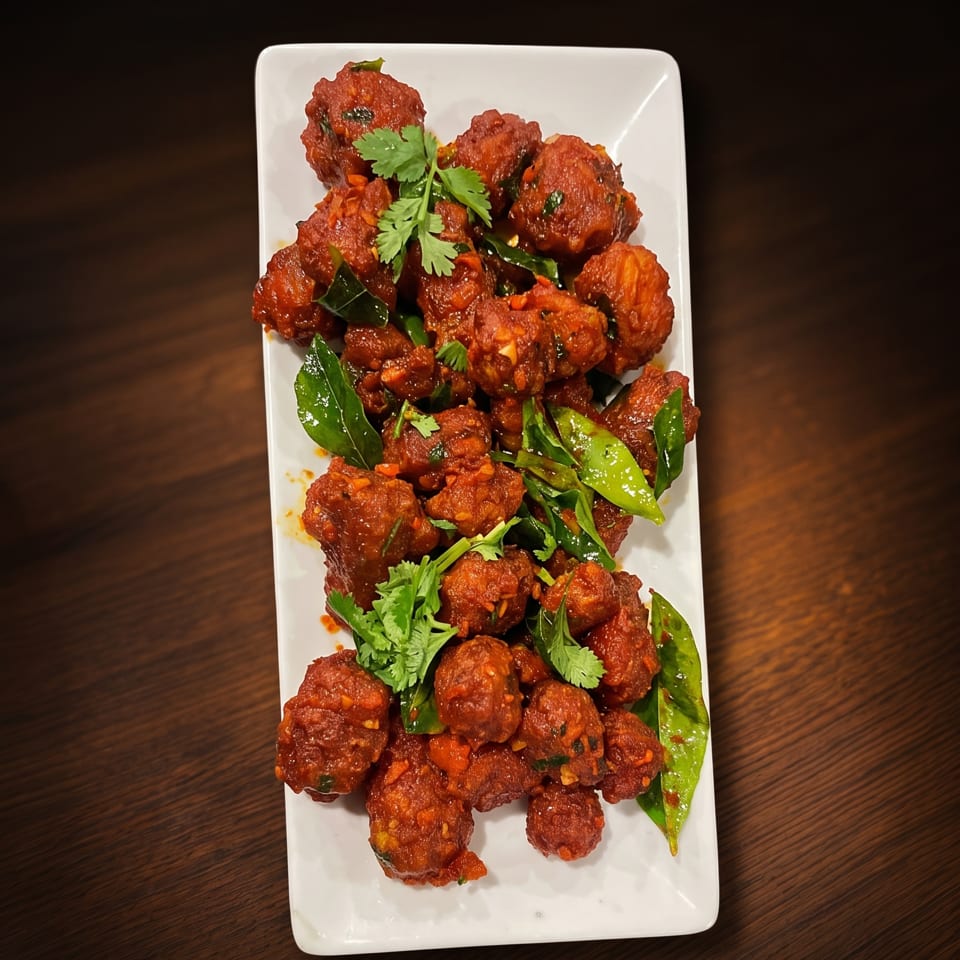 Spicy red fried chicken bites garnished with cilantro and curry leaves on a white plate.